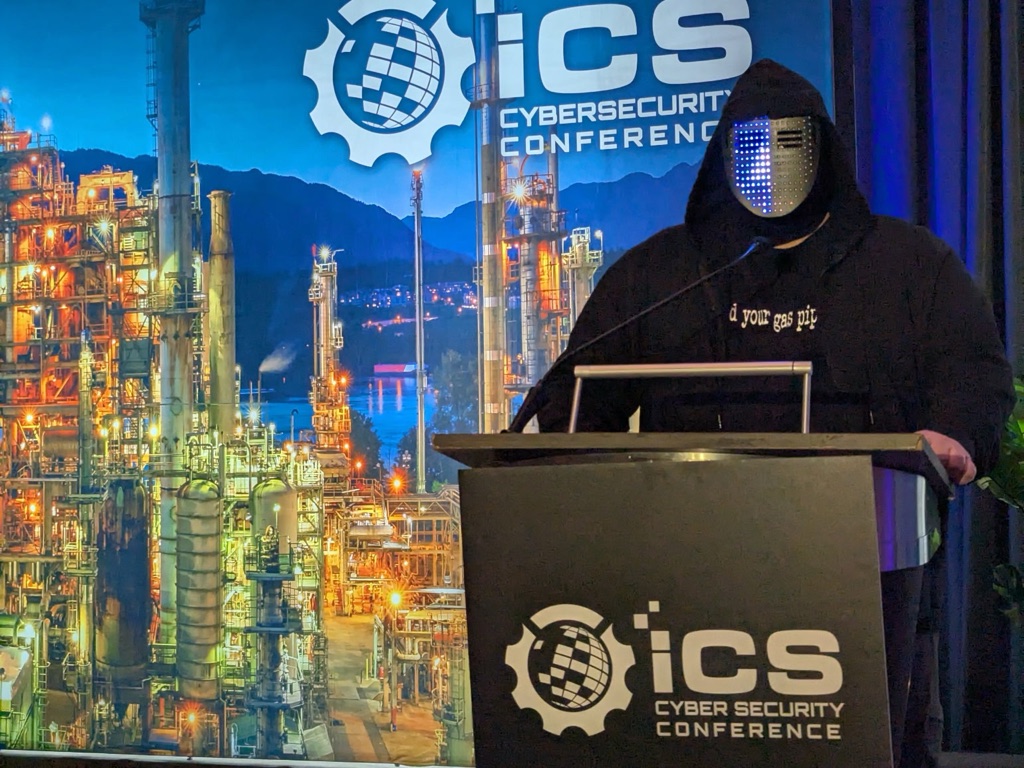 Clint Bodungen speaking at the podium in an LED mask at the ICS Cybersecurity Conference